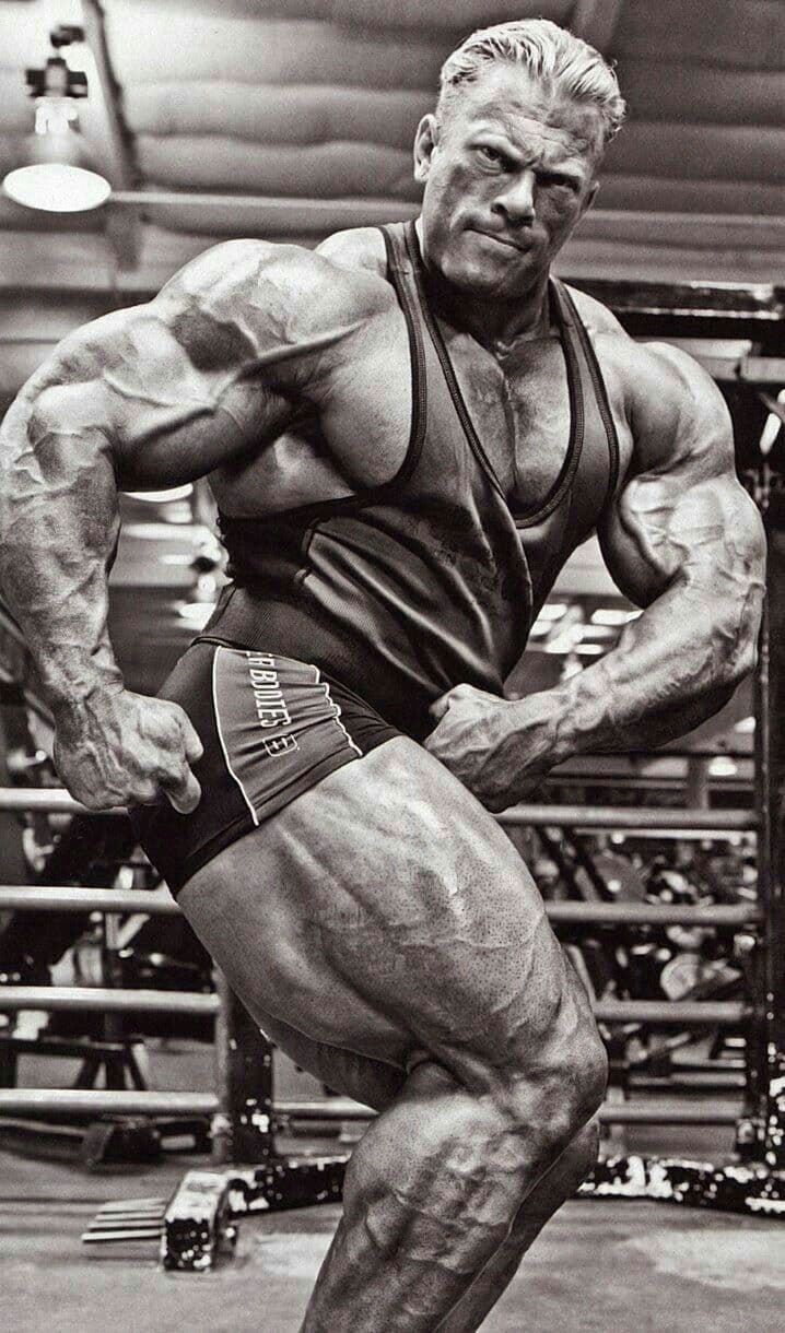 Ein muskulöser Bodybuilder posiert in einem Fitnessstudio, wobei er seine Arme und Beine anspannt, um seinen Körperbau zu betonen. Er trägt ein Tanktop und Shorts, und im Hintergrund sind Fitnessgeräte zu sehen. Das Bild ist schwarz-weiß.