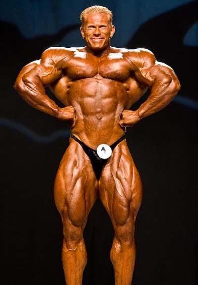 Ein männlicher Bodybuilder mit braungebranntem, muskulösem Körperbau posiert auf der Bühne in einem schwarzen Posierkoffer und lächelt selbstbewusst mit den Händen in den Hüften vor einem dunklen Hintergrund.