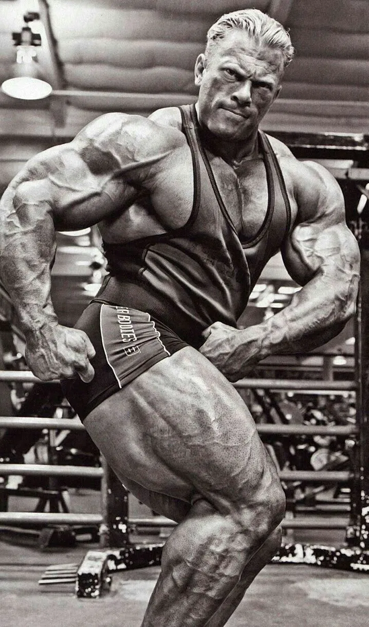 Ein muskulöser Bodybuilder posiert in einem Fitnessstudio, wobei er seine Arme und Beine anspannt, um seinen Körperbau zu betonen. Er trägt ein Tanktop und Shorts, und im Hintergrund sind Fitnessgeräte zu sehen. Das Bild ist schwarz-weiß.