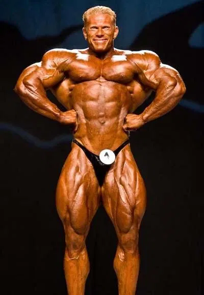 Ein männlicher Bodybuilder mit braungebranntem, muskulösem Körperbau posiert auf der Bühne in einem schwarzen Posierkoffer und lächelt selbstbewusst mit den Händen in den Hüften vor einem dunklen Hintergrund.