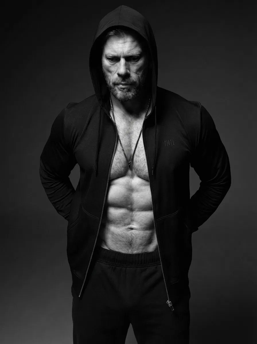 Ein muskulöser Mann mit Bart trägt eine Kapuzenjacke, die den Reißverschluss öffnet und seine Bauchmuskeln zum Vorschein bringt. Er steht mit den Händen auf dem Rücken vor einem dunklen, schlichten Hintergrund auf einem Schwarz-Weiß-Foto.