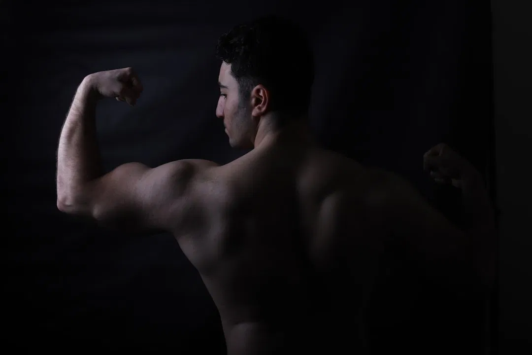 Eine hemdsärmelige Person lässt ihre Armmuskeln mit dem Rücken zur Kamera vor einem dunklen Hintergrund spielen, wodurch ihr muskulöser Körperbau hervorgehoben wird.