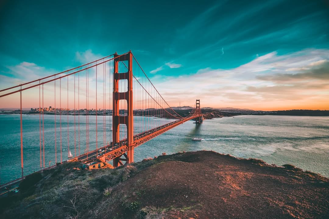 Die Golden Gate Bridge erstreckt sich bei Sonnenuntergang über das Wasser, ein Boot fährt darunter vorbei und die Skyline der Stadt ist in der Ferne unter einem dramatischen Himmel zu sehen.